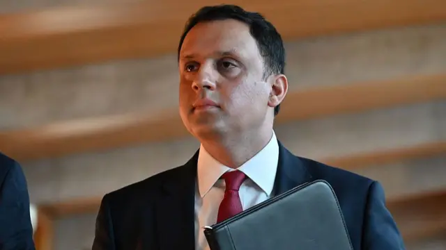 Anas Sarwar wearing a dark suit, white shirt and red tie and holding a folder