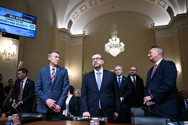 Three men in dark suits stand behind a table in a large room with a vaulted ceiling and chandelier
