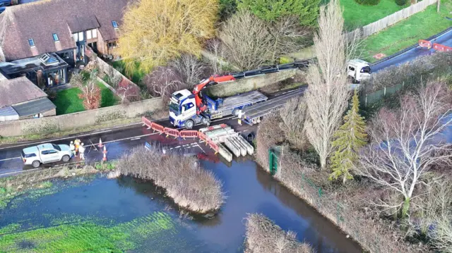 A drone image of a flooded junction. Water covers a huge area of road. The road is fenced off and there are two lorries dealing with the flood