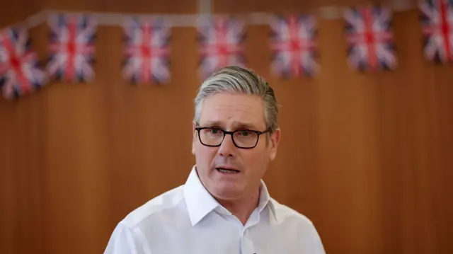 Keir Starmer with his right hand in a fist as he gestures while speaking to a room of people sitting in circle. He's in a white shirt, the background is blurred but there's Union flag bunting visible on the wall