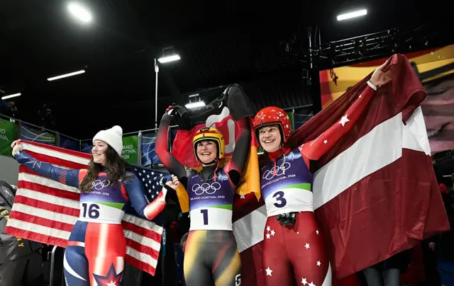 Third-placed USA's Ashley Farquharson, first-placed Germany's Julia Taubitz and second-placed Latvia's Elina Bota celebrate withe their national flags after crossing the finish line in the luge women's singles run 4 at Cortina Sliding Centre during the Milano Cortina 2026 Winter Olympic Games in Cortina d'Ampezzo