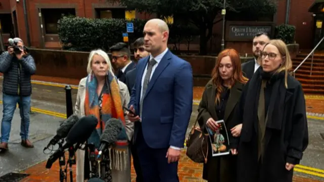 A bald man in a blue suit, surrounded by three women in coats, standing in the street in front of microphones on a tripod