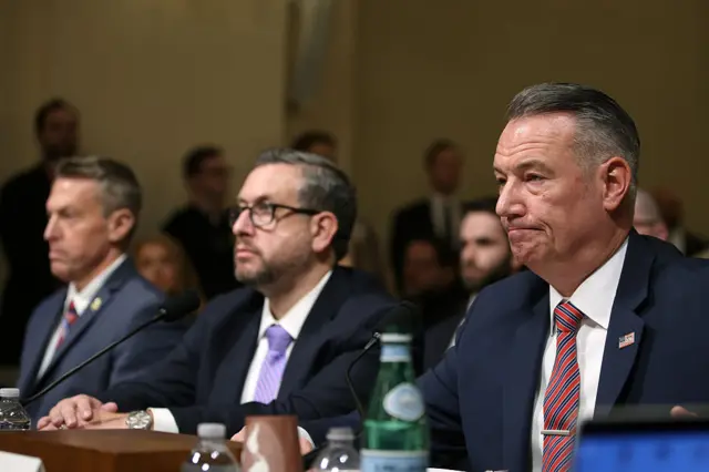 Three men in suits and ties sit at a desk behind microphones