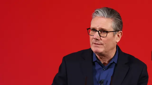 Keir Starmer in a black suit jacket and dark blue shirt speaking in front of a red wall