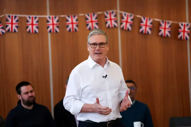 Keir Starmer in white shirt standing in the centre of a room surrounded by people sitting down