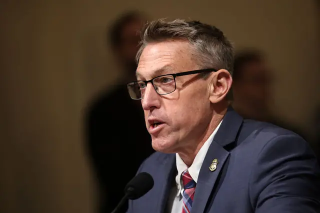 A man with ligth brown hair, black-framed glasses and blueish grey suit speaks into a microphone