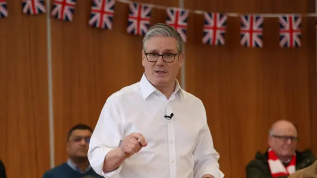 Starmer speaking to a small audience in a community. He's wearing a white shirt without a tie. Behind him are lots of union jack flags hung up in bunting.