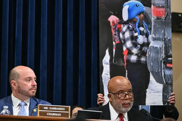Two bald men sit behind a desk with a large photo of a child in a blue hat behind them