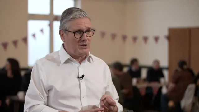 Keir Starmer in white shirt answering questions. Behind him, in the blurred background, are people sitting down around wooden table, Union flag bunting hanging from the walls