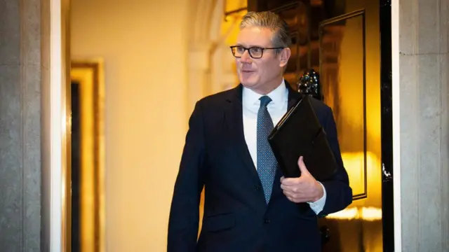 Keir Starmer walking out the door of 10 Downing Street holding a black folder. It is dark outside.
