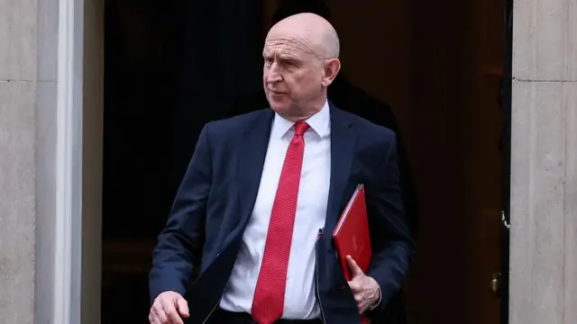 Defence Secretary John Healey wears a dark suit with a bright red tie. He is carrying a slim red folder