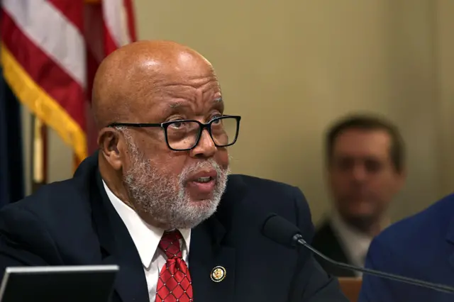 A bald man with a white beard, black glasses and a red tie speaks into a microphone