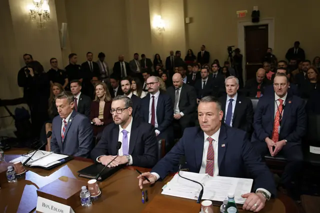Three men in suits sit behind a desk with microphones, paper and water bottles in front of them
