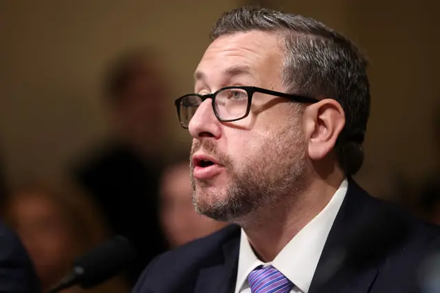A man with greying hair, a beard and glasses speaks into a microphone