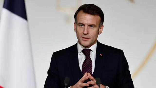 Emmanuel Macron in a dark suit and red dotted tie, he is standing behind a podium with his hands together, he has dark hair.