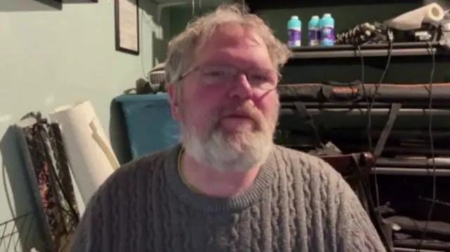 Ernst Trent, a white man with white hair and beard, wearing a brown cable knit jumper say on a chair.