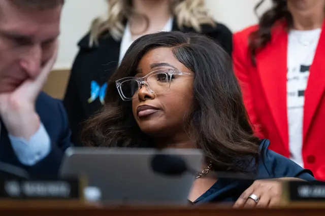 A woman behind a desk with black hair and clear-rimmed glasses looks to her right