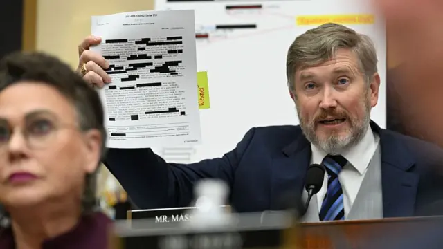 Massie sits behind the dais and speaks into a mic. He holds up a piece of paper with two paragraphs of text on it. Many words are redacted. The document has a sticky note on it labeled "Wexner".
