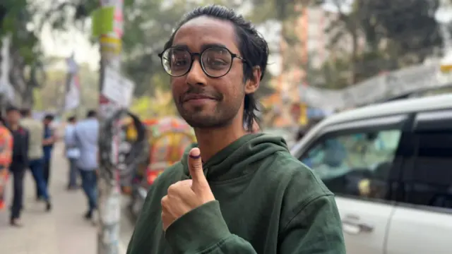 A man wearing classes and a green sweatshirt gives a thumbs up