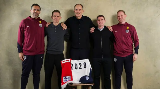 Thomas Tuchel poses with his staff behind an England shirt with '2028' on the back.