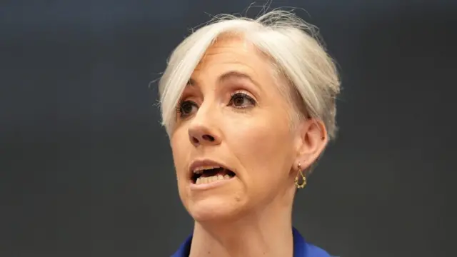 Daisy Cooper, a woman with short blonde hair and a blue jacket, speaks against a dark background.