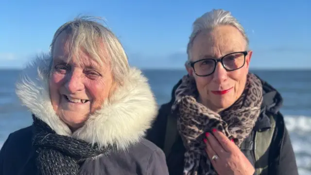 Two women in winter coats stood by the sea