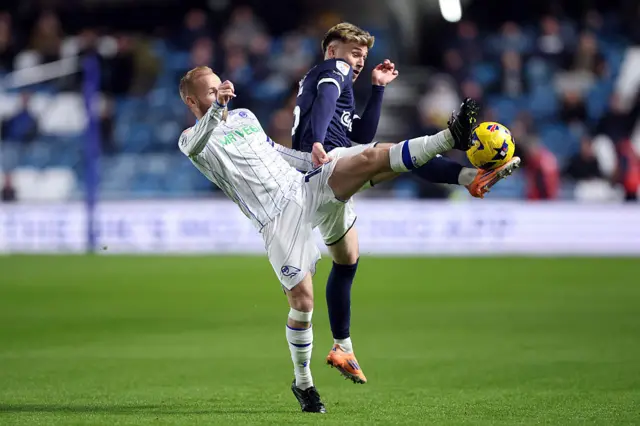 Barry Bannan, dressed in Sheffield Wednesday's white away kit with blue pinstripes, and Luke Cundle, wearing Millwall's home kit of a dark blue top, white shorts and blue socks, challenge each other for the ball, at waist height with both players putting their leg up and making contact with the ball