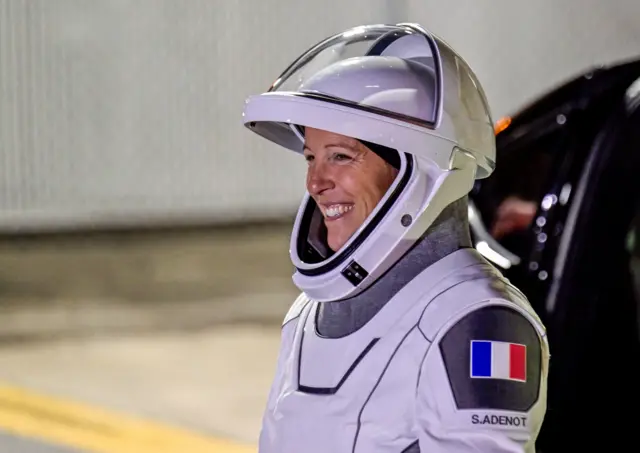 Sophie Adenot walks in her white space suit which has a French flag on the shoulder - she is smiling