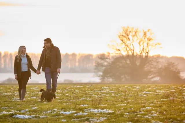 A couple holding hands walking their dog in a park with snow and frost on the grass.
