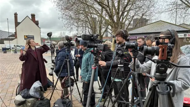 Multiple journalists with cameras gather outside a courtroom.