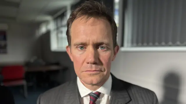 A man looking into the camera wearing a grey suit and a tie.