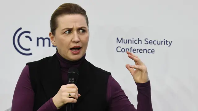 Danish Prime Minister Mette Fredericksen mid-speech, a mic in her right hand as she gestures with her left one. She's in a black button-up vest and purple crew neck shirt