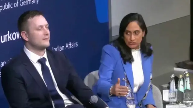 A man in a suit and tie sat beside a woman in a blue suit, speaking while gesturing her hand