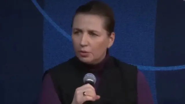 Danish Prime Minister Mette Frederiksen speaking into a microphone at a press conference. She is wearing a purple long sleeved top with a black polo neck jumper