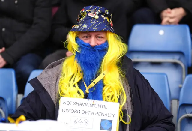 An Oxford United fan wearing a fake beard and a sign with the text 'Not Shaving Until Oxford Get A Penalty' before the Emirates FA Cup fourth round match at Kassam Stadium, Oxford.