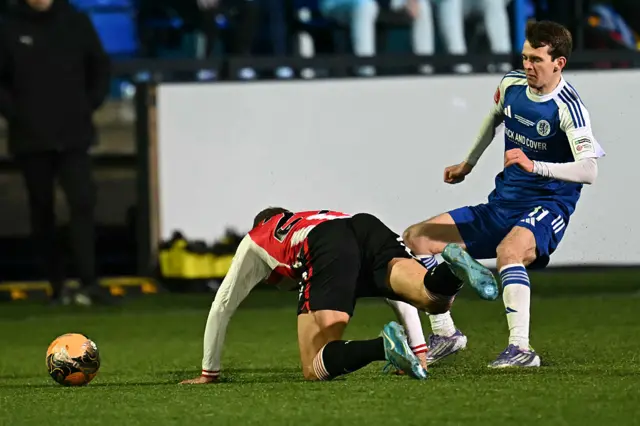 Brentford's German midfielder #27 Vitaly Janelt (L) is booked for this foul on Macclesfield's English striker #11 Luke Duffy (R) during the English FA Cup fourth round football match between Macclesfield Town and Brentford at Leasing.com Stadium, Moss Rose in Macclesfield, northern England on February 16, 2026.