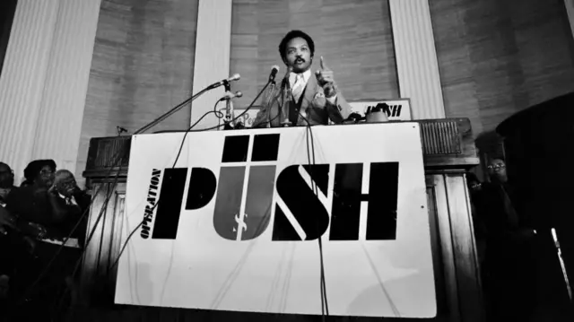 A black-and-white photo of Jackson speaking at a podium. There's a big banner below him which says 'PUSH'