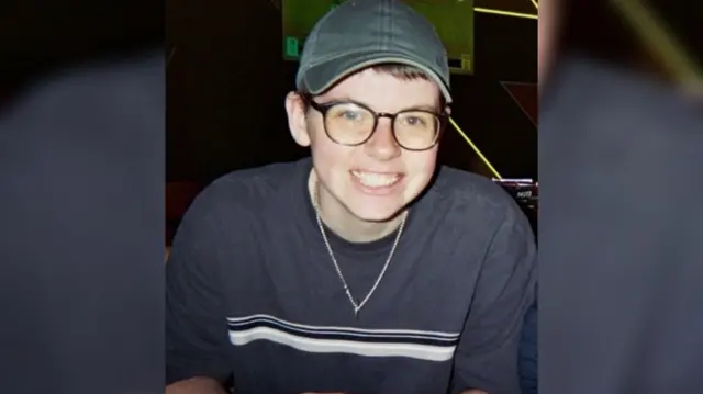 A man smiling in front of a camera. He's wearing glasses and a green cap