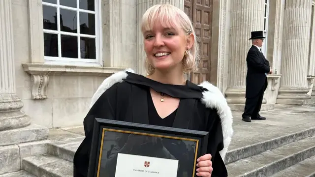 A medium close up of a smiling Lucy Gabb wearing her graduation gown and holding her framed Cambridge University degree outside of a Georgian stone building.