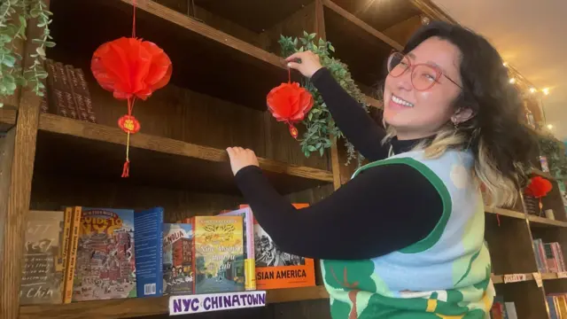 A woman wearing glasses hangs a red lantern on a bookcase