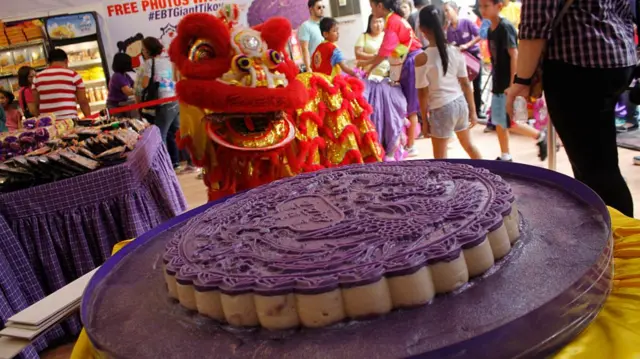 A giant sticky rice cake topped with purple yam frosting with a dragon dancer in the background
