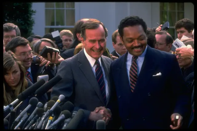 Jesse Jackson and George Bush pictured in front of a crowd of reporters with microphones in November 1988