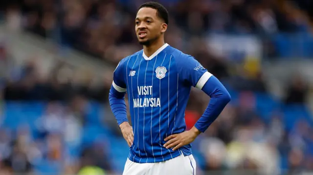Chris Willock stands with his hands on his hips, wearing Cardiff City's blue jersey with white pinstripes and white short
