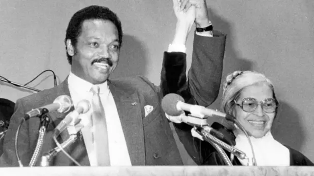 A black-and-white image of Jesse Jackson Sr and activist Rosa Parks raising their hands triumphantly during a speech, 1965.