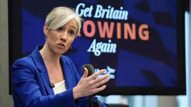 Daisy Cooper speaking at an event. Behind her is a television screen which reads 'Get Britain Growing Again'.