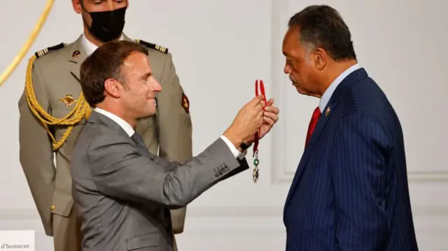 Macron standing with Jackson, both in suits. Macron is placing a medal of honour around Jackson's neck