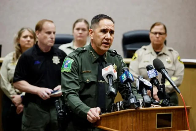 A man in a green law enforcement uniform stands at a bank of microphones