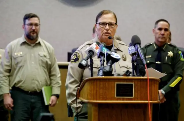 A woman in a sheriff's uniform stands behind a lectern covered in microphones, two men in green uniforms stand behind her