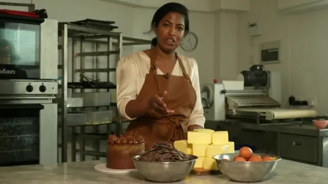 Gaya Vara of Gaya Bakery in London stood in her bakery's kitchen wearing a brown airport with a white blouse underneath gesturing at a chcolate cake, a bowl of chocolate, a stack of butter, and a bowl of eggs in front of her on a table