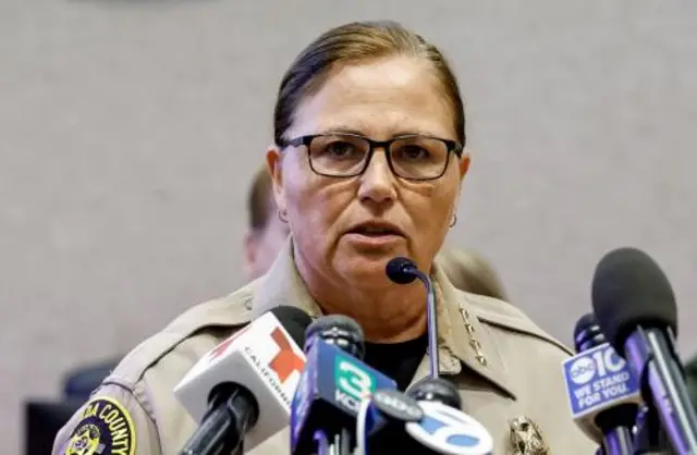 A woman in a sheriff's uniform and glasses stands behind a bank of microphones
